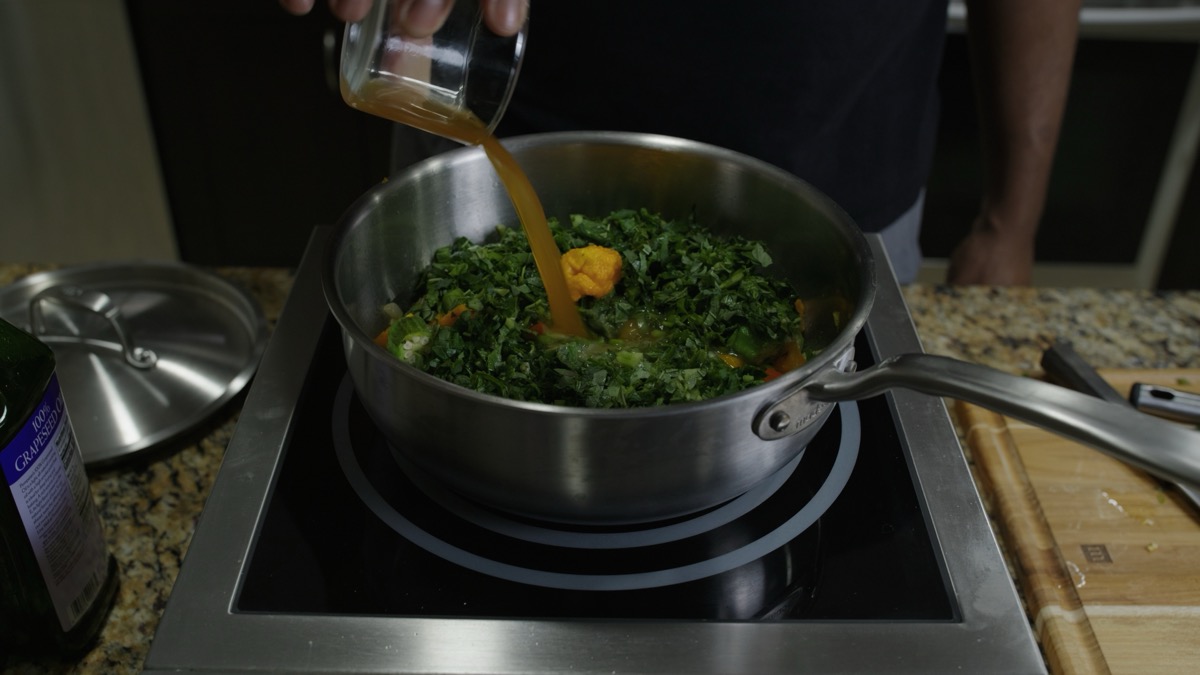 Pouring vegetable stock over chopped callaloo in the pot